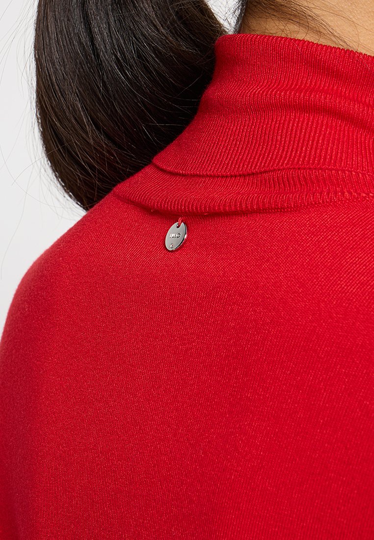 Maglione a collo alto rosso con texture a coste e un'etichetta d'argento rotonda nella parte posteriore del collo. Il tessuto sembra leggero e liscio.