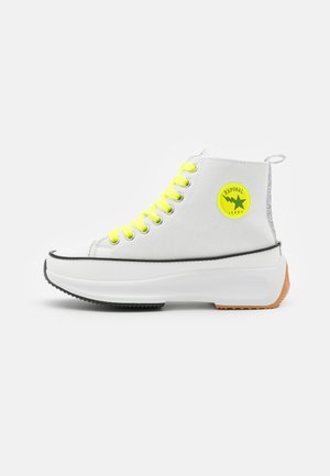 Hoge sneakers van wit canvas met felgele veters, zwarte rubberen bies en een cirkelvormig groen logo. Dikke witte zool met een tan onderkant.