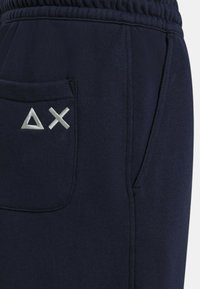 Pantaloni sportivi blu navy realizzati in tessuto morbido, con tasca laterale e ricamo di simboli triangolari bianchi e una X vicino alla tasca. Vita elasticizzata.