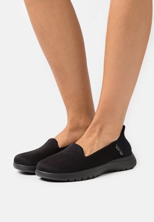 Black slip-on shoes with a textured upper, rounded toe, and soft sole. Features a logo accent on the side and elasticated entry for comfort.