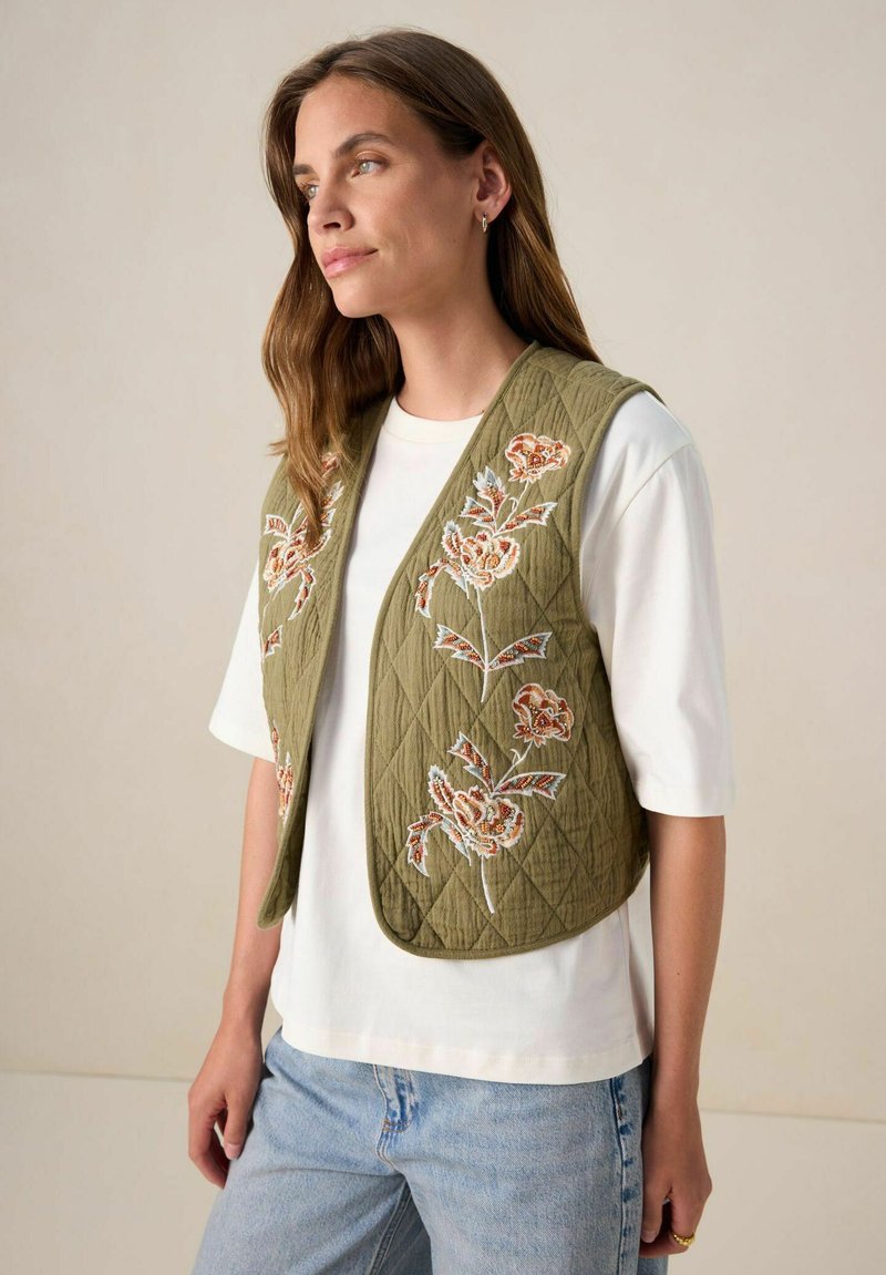 Gilet en olive matelassé avec broderies florales en rouge et bleu. Associé à un t-shirt blanc, le look est complété par un jean en denim clair.