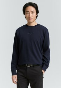Marineblauw sweatshirt met lange mouwen, voorzien van een geribbelde ronde hals en een klein, donker logo op de borst. Gepaarde met donkere broek en een riem.