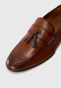 Mocasín de cuero marrón con borlas de color marrón oscuro y detalle de costura sobre fondo blanco, mostrando ángulos frontal y lateral.