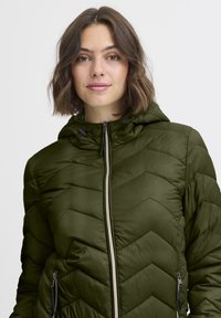 Karinė žalios spalvos pūkinė striukė su chevron raštu, priekyje užtrauktuku užsegama ir su reguliuojama kapišonu. Turi šonines kišenes su užtrauktukais.