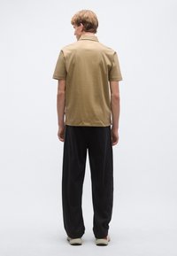 Svetlohnedá košeľa s krátkym rukávom a golierom, v kombinácii s tmavými voľnými nohavicami. Topánky typu Vans so svetlými a červenými akcentmi. Jednoduché pozadie.