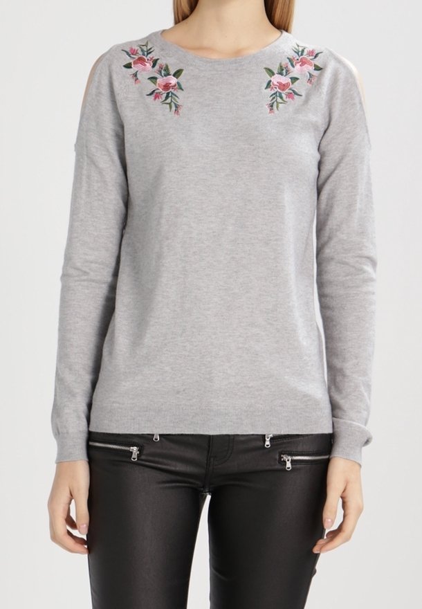 Grå långärmad tröja med blommig broderi på axlarna, med öppna axelutskärningar; ihop med svarta byxor med dragkedjedetaljer.