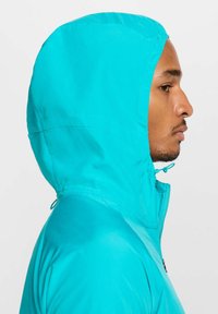 Turquoise capuchonjack met ritssluiting, gladde stof en verstelbare trekkoord bij de hals, gezien vanaf de zijkant.