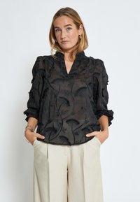 Blusa nera trasparente con ricami floreali, scollo a V e maniche a sbuffo. Presenta un tessuto testurizzato e delicati dettagli a frill.