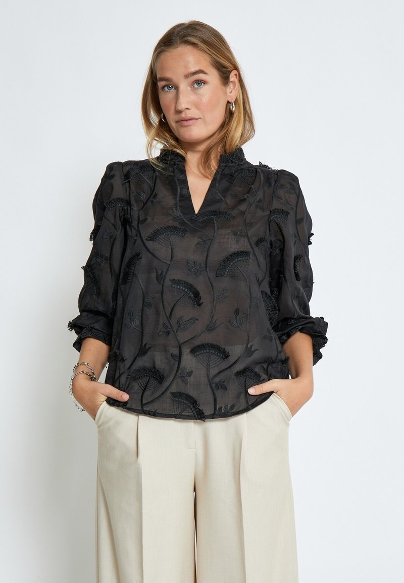 Blusa nera trasparente con ricami floreali, scollo a V e maniche a sbuffo. Presenta un tessuto testurizzato e delicati dettagli a frill.