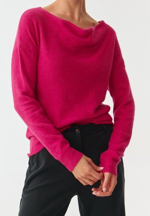 Kobieta w jasnoróżowym, długim swetrze z szerokim dekoltem i czarnych spodniach, z lekko złożonymi rękami na talii.