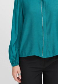 Blusa teal con maniche lunghe, polsini arricciati e una cerniera frontale con dettagli metallici. Abbinata a pantaloni neri con una vestibilità rilassata.