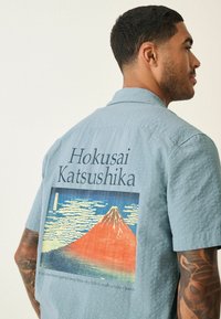 Svijetloplava košulja kratkih rukava s teksturiranim materijalom. Na leđima je ilustracija planine Fuji s oblacima i crnim natpisom.