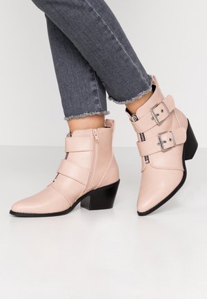 Bottines beige à talons carrés noirs, avec double boucle et fermetures éclair, portées avec un jean skinny gris à ourlet effiloché.