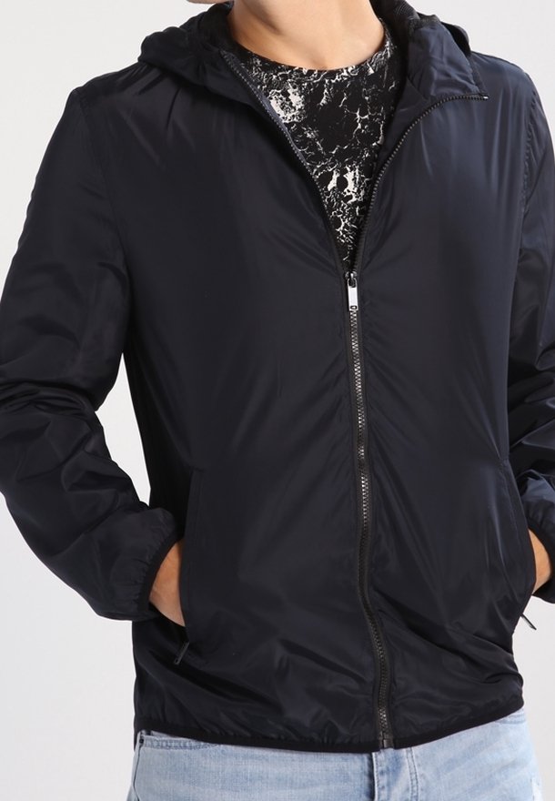 Svart lättviktsjacka med huva och dragkedja framtill, med elastiska ärmar och en jämn textur. Bärs över en mönstrad skjorta.