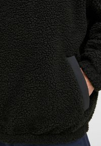 Black fleece pullover made of textured fabric, featuring a pocket with a contrasting trim. The design is relaxed-fit with elasticated cuffs.
