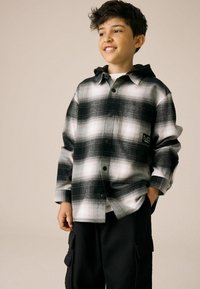 Chemise en flanelle à carreaux noir et blanc avec une capuche, ornée de grands boutons et d'une poche sur la poitrine. Associée à un pantalon cargo noir.