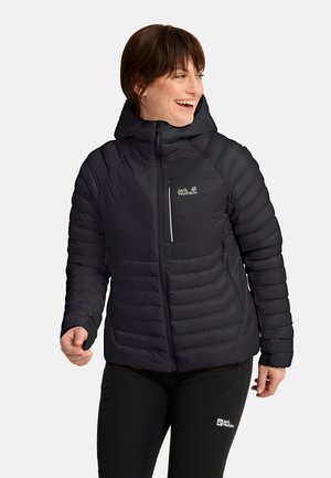 Donna che indossa una giacca a vento nera con cappuccio e pantaloni neri con loghi Jack Wolfskin, sorridente e che guarda di lato.