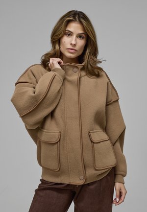 Femme aux cheveux mi-longs portant une veste polaire marron oversize avec de grandes poches avant, debout devant un fond gris.