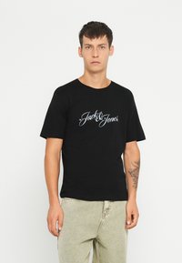 Fekete pamut póló rövid ujjakkal, középen fehér írásos "Jack & Jones" logóval a mellkason. A modellen világos színű nadrág van.