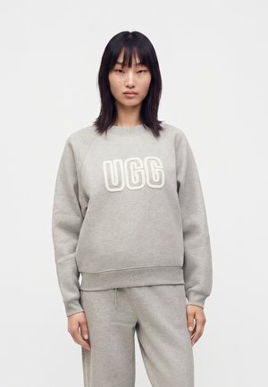 Joven mujer con cabello negro liso lleva una sudadera gris UGG y pantalones deportivos a juego sobre un fondo blanco liso.
