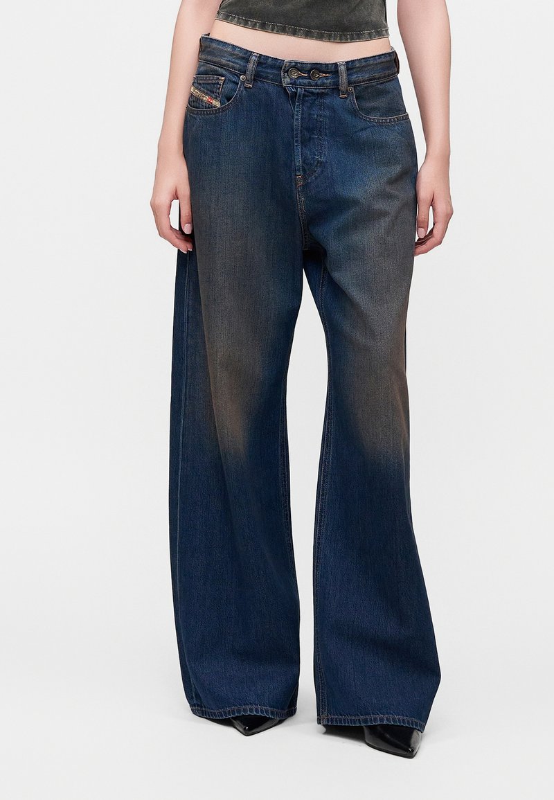 Personne portant un jean en denim bleu foncé à jambes larges et un haut noir raccourci, debout devant un fond clair uni.