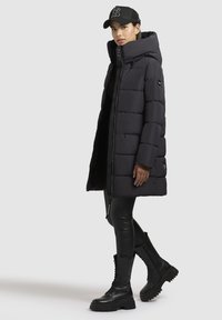 Zwarte pufferjas met hoge kraag en ritssluiting, gecombineerd met zwarte leren leggings en combat boots, aangevuld met een zwarte pet.