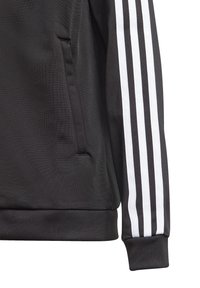 Musta takin hiha, jossa on kolme rinnakkaista valkoista raitaa ja sivutasku, esittelee ribbikudottua hihansuut ja helman yksityiskohtia.