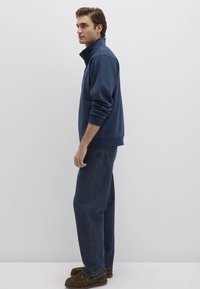 Dunkelblauer Pullover mit hohem Kragen, gerippten Bündchen und Saum. Kombiniert mit geraden, dunklen Jeans und braunen Slippers.