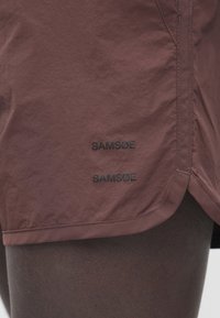 Krótkie brązowe sportowe szorty wykonane z lekkiej tkaniny, z dwoma czarnymi logo "SAMSØE" w pobliżu brzegu oraz prostym, funkcjonalnym designem.