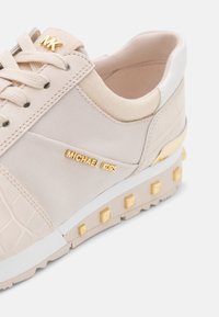 Baskets beige Michael Kors en tissu texturé, détails du logo doré sur le côté et les lacets, semelle blanche et clous carrés dorés sur la semelle intermédiaire.