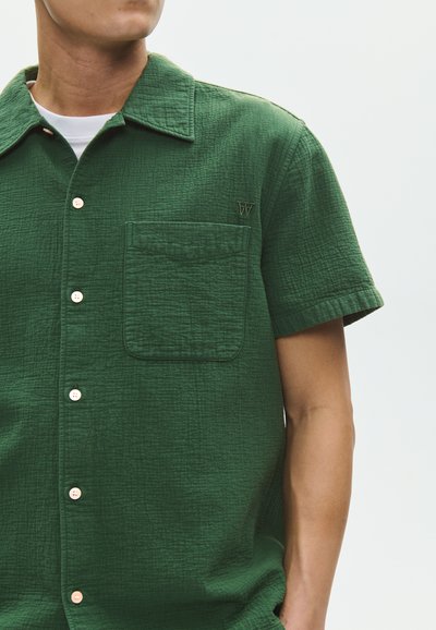 Homme portant une chemise boutonnée texturée à manches courtes vert foncé avec une poche poitrine et un tee-shirt blanc en dessous, sur un fond uni.