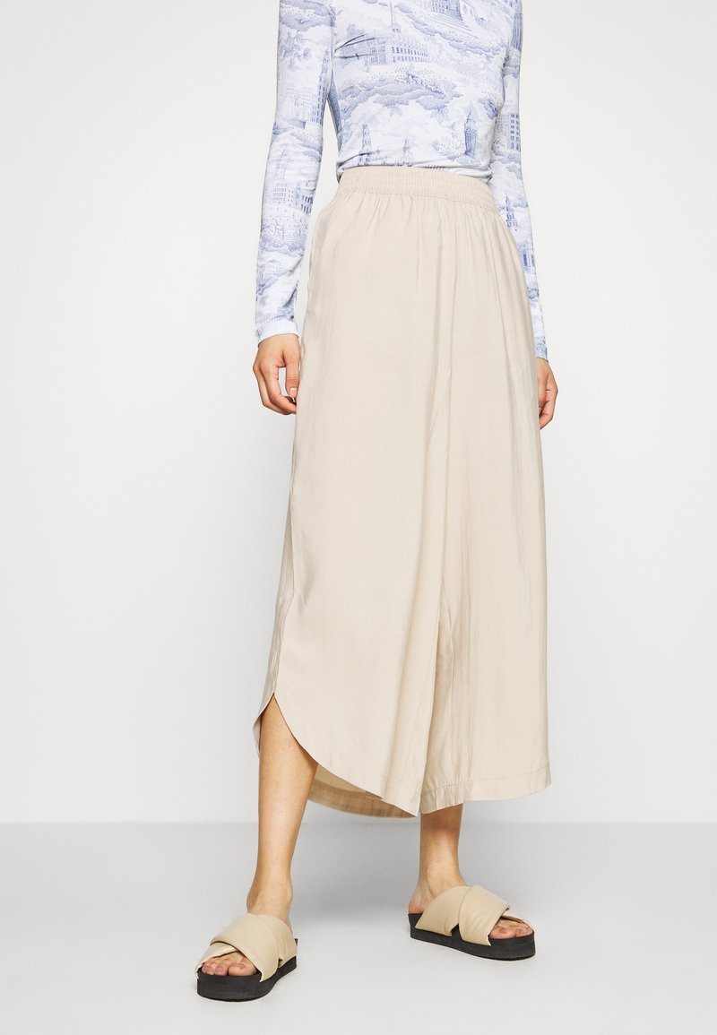Femme portant une jupe midi beige taille haute avec un ourlet asymétrique, associée à des sandales beiges à enfiler et un haut imprimé à manches longues.