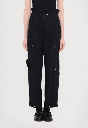 Czarne spodnie cargo z wysokim stanem i pasem typu paperbag, liczne kieszenie z patkami, złote guziki, noszone z czarnymi loafersami i czarną bluzką włożoną do środka.