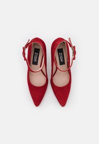 Bottes à talons hauts en suede rouge avec un bout pointu et une bride de cheville. Texture lisse et côtés ouverts. Logo de la marque visible sur la semelle intérieure.