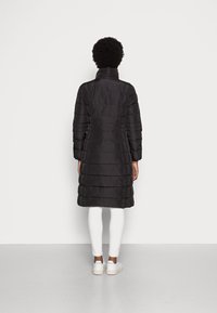 Cappotto imbottito nero con colletto alto, caratterizzato da sezioni trapuntate orizzontali, maniche lunghe e design di lunghezza intermedia. Indossato sopra pantaloni bianchi.