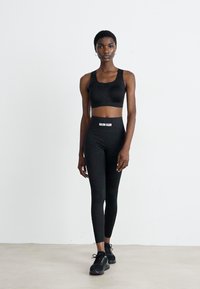 Žena v černém sportovním podprsenka, s legínami vysokého pasu s logem Calvin Klein a černými teniskami, stojící proti pozadí bílé zdi.