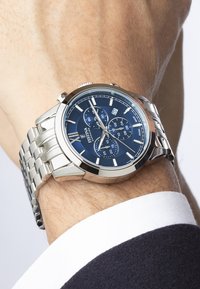 Reloj de acero inoxidable plateado con esfera azul, que presenta funciones de cronógrafo, manecillas plateadas y una pulsera de metal cepillado.