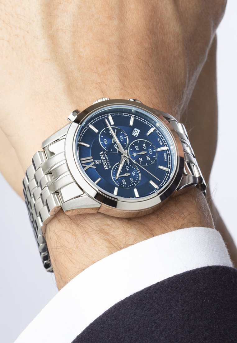 Reloj de acero inoxidable plateado con esfera azul, que presenta funciones de cronógrafo, manecillas plateadas y una pulsera de metal cepillado.