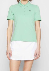 Polo shirt verde claro de algodón, con cuello, mangas cortas y un pequeño logo de Lacoste en el pecho. Combinado con una falda blanca.