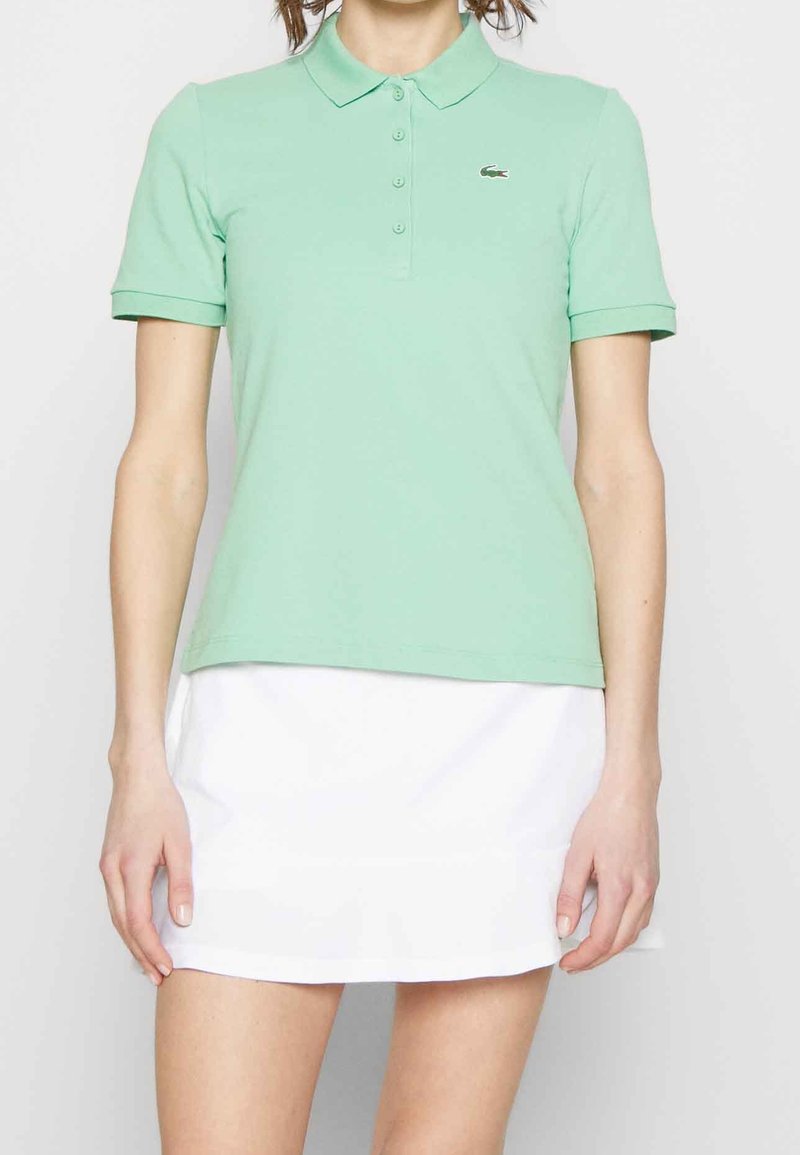 Polo shirt verde claro de algodón, con cuello, mangas cortas y un pequeño logo de Lacoste en el pecho. Combinado con una falda blanca.
