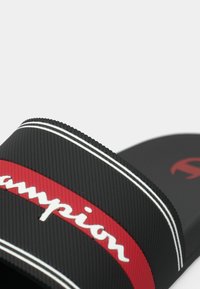 Μαύρη λαστιχένια σαγιονάρα με υφασμένη λουρίδα, κόκκινη ταινία και λευκό λογότυπο, διακοσμημένη με λευκές ρίγες. Ελαφρύ και ανθεκτικό σχέδιο.