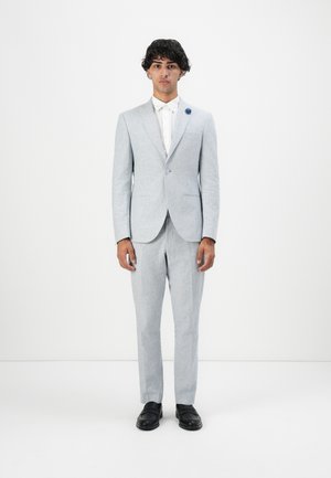 Νεαρός άνδρας με ανοιχτό γκρι κοστούμι, λευκό πουκάμισο, μαύρα παπούτσια και μπλε καρφίτσα με φόντο λευκό.