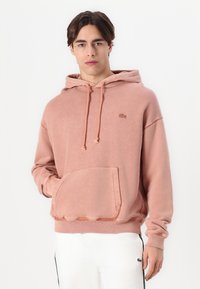 Sudadera con capucha rosa, con bolsillo tipo canguro, puños acanalados y un pequeño logo en el pecho. Textura suave y diseño casual.
