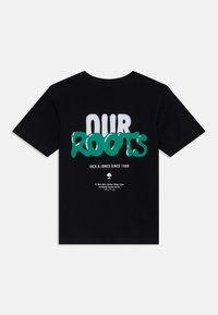 T-shirt en coton noir avec un texte graphique vert "NOS RACINES" sur un fond blanc décoloré. Inclut le nom de la marque et des détails imprimés en petit en bas.