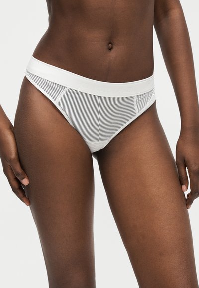 Femme portant un sous-vêtement style bikini blanc avec de fines rayures verticales noires et une ceinture blanche unie, sur un fond uni.