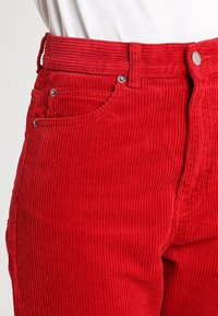 Pantaloni in corduroy rossi con righe verticali, dotati di una vita alta, cinque tasche e dettagli in metallo argentato.