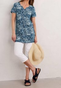 Túnica de manga corta con estampado floral azul, combinada con pantalones cortos blancos y sandalias negras informales, sosteniendo un sombrero de paja marrón.