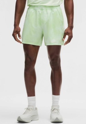 Mann trägt hellgrüne Shorts, weiße Socken und weiße Laufschuhe, steht vor einem schlichten weißen Hintergrund.