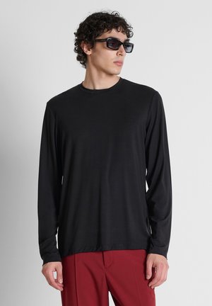 Uomo con capelli ricci che indossa una camicia nera a maniche lunghe, pantaloni rossi e occhiali da sole neri, che guarda di lato su uno sfondo grigio uniforme.