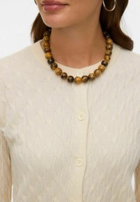 Kremowy kardigan z dzianiny w sploty warkoczowe, z okrągłym dekoltem i pięcioma guzikami. Noszony jest wielokolorowy naszyjnik z koralików w brązowych i złotych odcieniach.